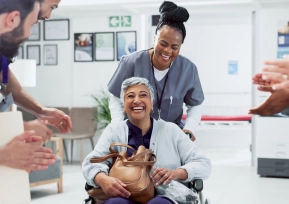 Profissionais de saúde aplaudem paciente sorridente em cadeira de rodas durante atendimento humanizado.