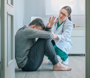 Jovem angustiado sentado no chão, com a cabeça baixa, enquanto uma profissional de saúde, vestindo jaleco, agacha-se ao lado dele com uma expressão de apoio e preocupação. A imagem representa o tema da saúde mental e o apoio psicológico.