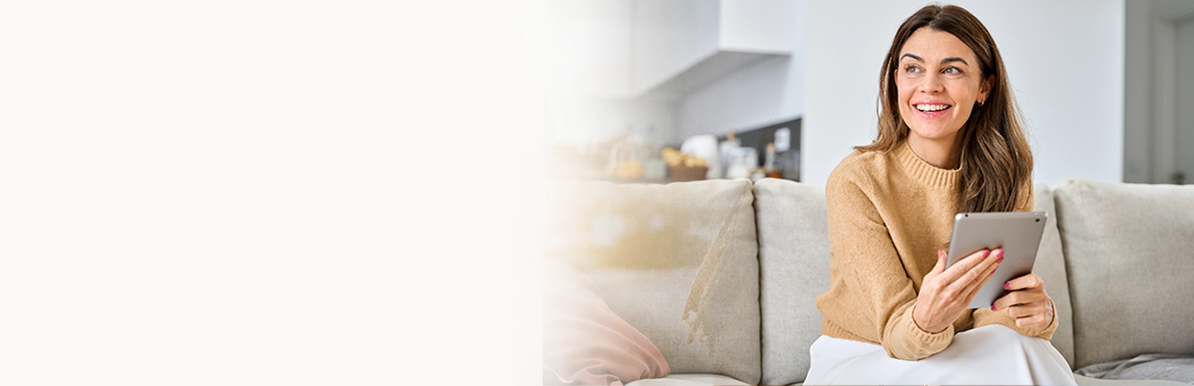 Mulher sorrindo e usando tablet em ambiente residencial moderno e bem iluminado, com sofá claro e cozinha ao fundo. Imagem representa uso de tecnologia em casa, conforto, conectividade e estilo de vida digital.