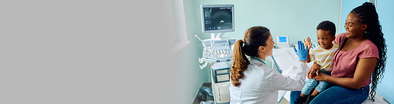 Médica interage com uma criança e um adulto em um consultório. A criança está sentada no colo do adulto, ambos sobre uma maca ambos sorrindo.