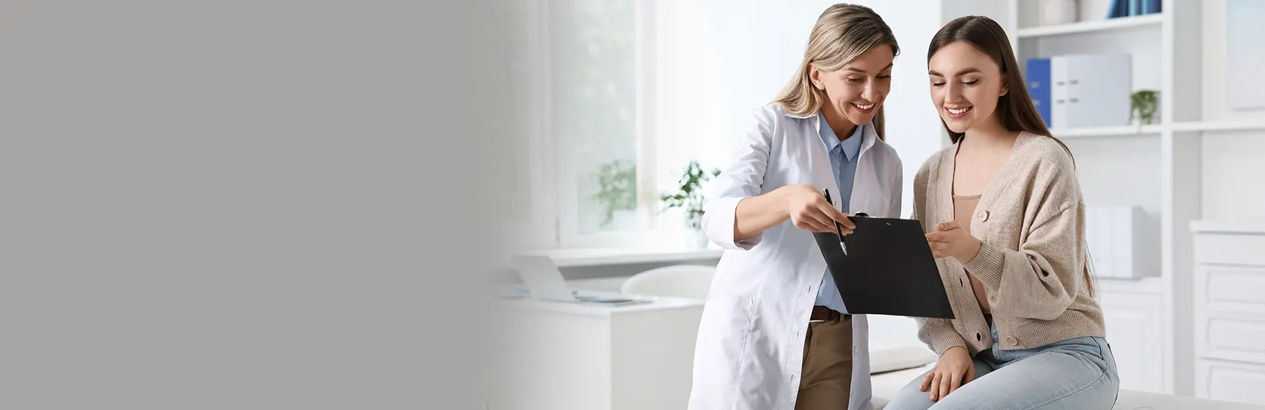 Profissional de saúde com jaleco branco e prancheta aponta informações para paciente sentada em maca, vestindo cardigã bege e jeans. Ambos sorriem em ambiente clínico moderno com janelas amplas, luz natural e decoração com livros e plantas ao fundo.