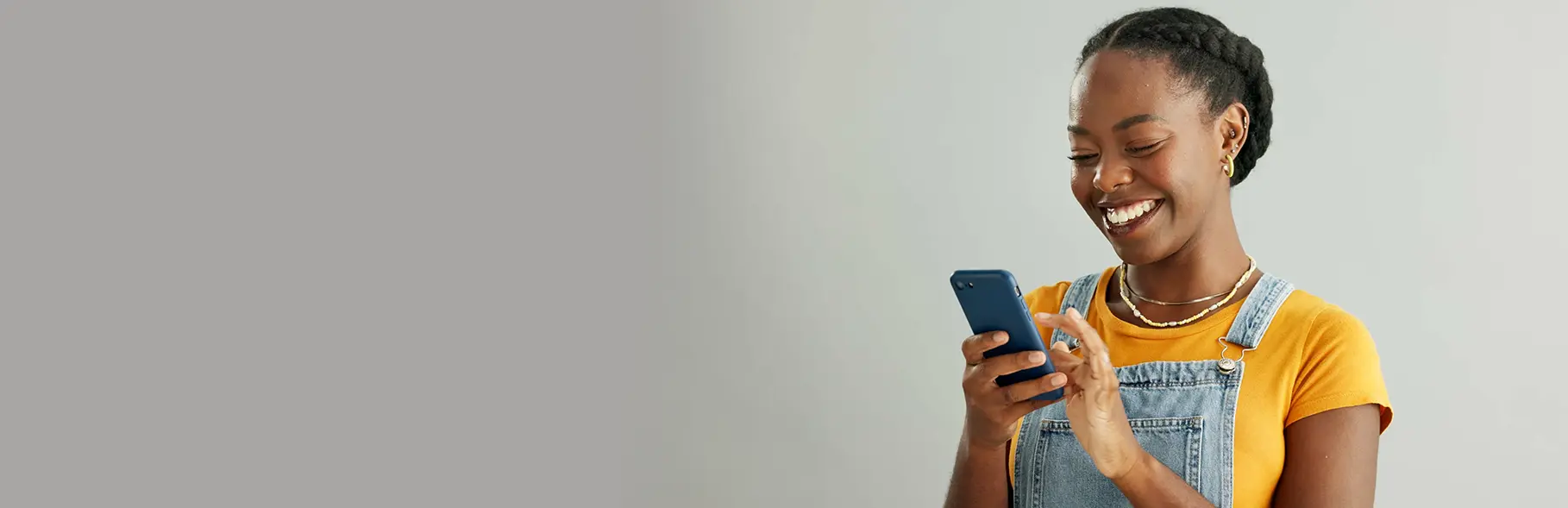 Mulher com camisa amarela e jardineira jeans segurando um smartphone e sorrindo, em ambiente interno.