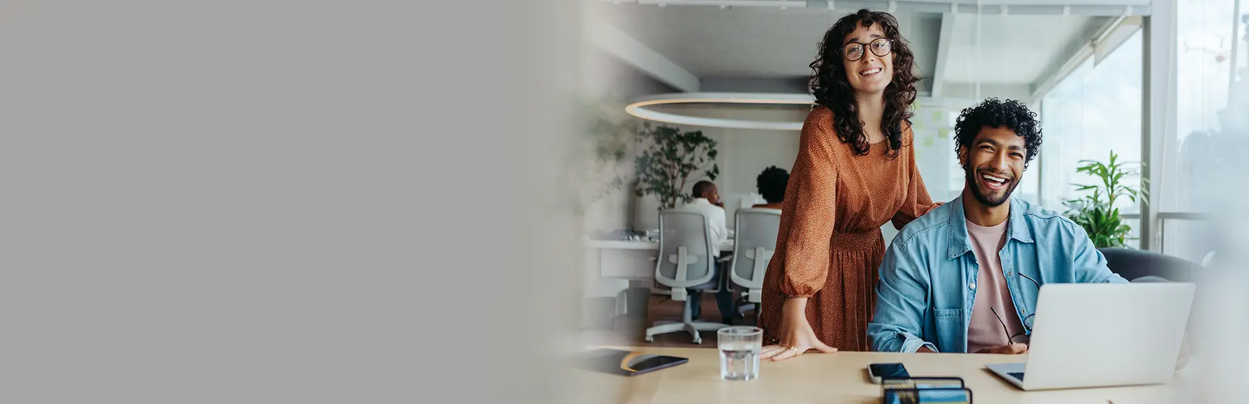 Homem sentado à mesa em ambiente de escritório, sorrindo enquanto trabalha em um notebook e uma mulher em pé ao lado sorrindo.