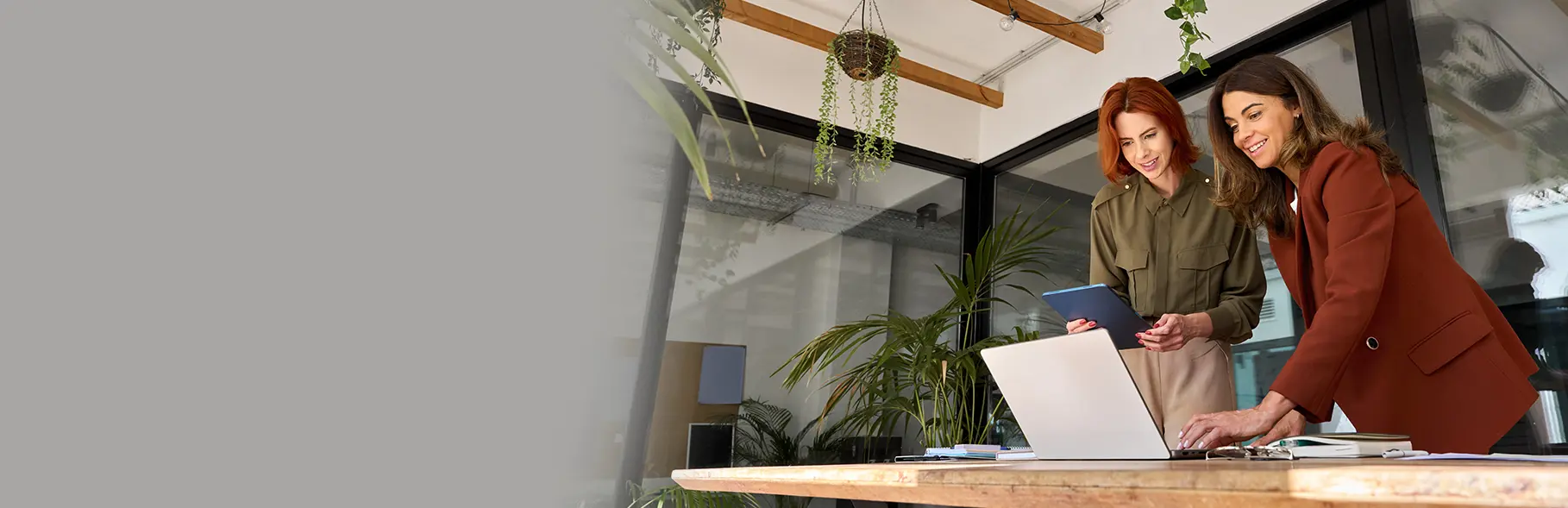 Duas mulheres em pé diante de uma mesa de madeira em ambiente de escritório moderno, uma está segurando um tablet enquanto a outra interage com um notebook, sugerindo colaboração em tarefas profissionais.