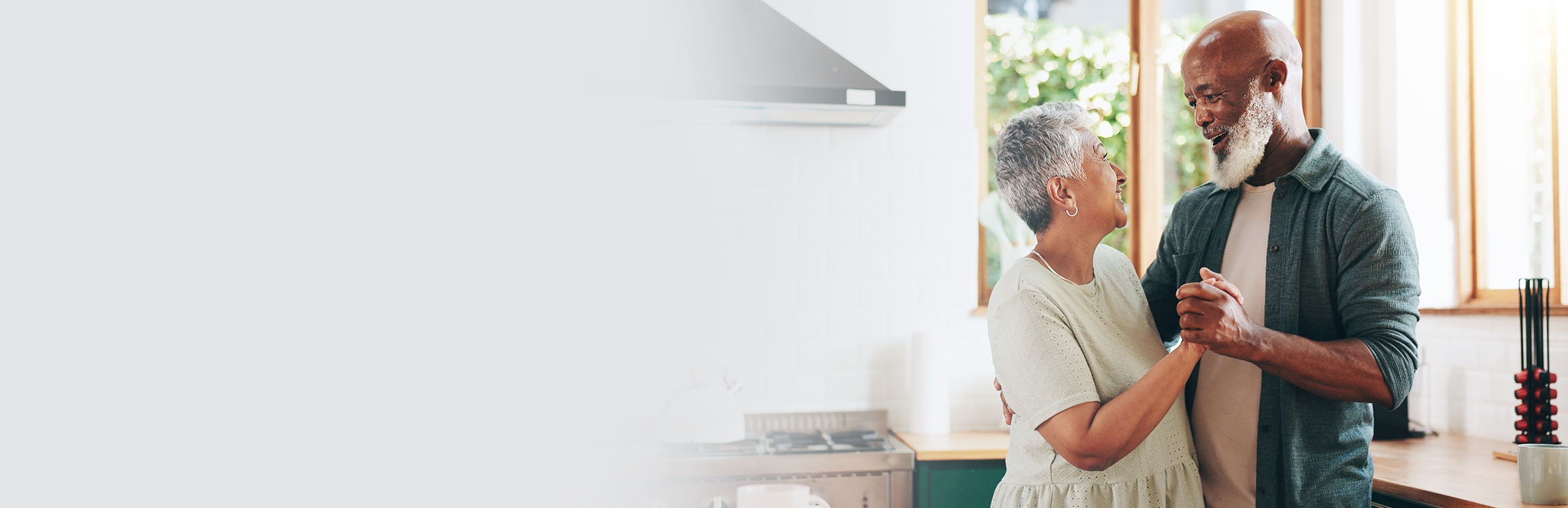 Um casal de idosos dançando juntos em uma cozinha, com fogão, bancada e janelas ao fundo mostrando vegetação externa.