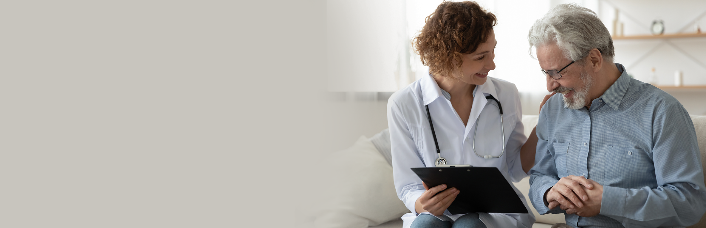 Médica com jaleco branco e estetoscópio, sentado ao lado de pessoa com óculos em sofá, segurando uma prancheta durante consulta.