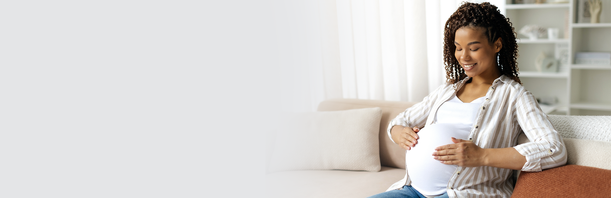 Mulher com cabelo cacheado sentada em sofá com fundo claro, vestindo camisa listrada sobre blusa branca, com as mãos sobre a barriga, em ambiente interno com iluminação suave.