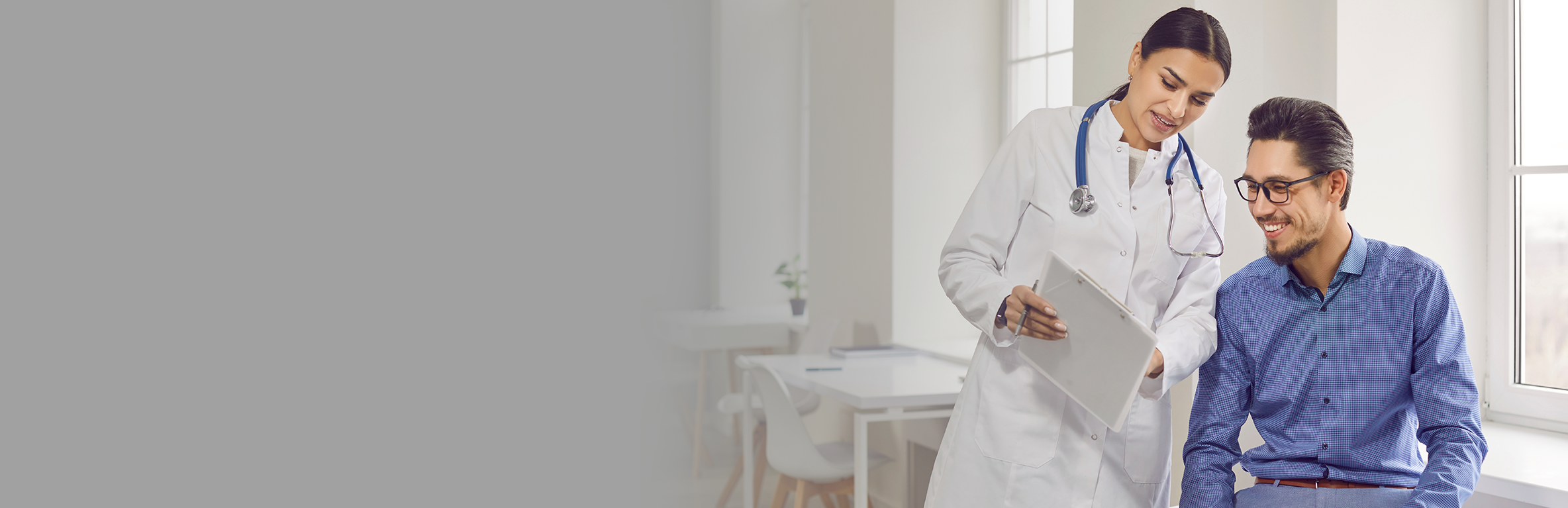 Médica vestindo jaleco branco segurando uma prancheta, conversando com o paciente de óculos, sentado próximo à janela, em ambiente clínico.