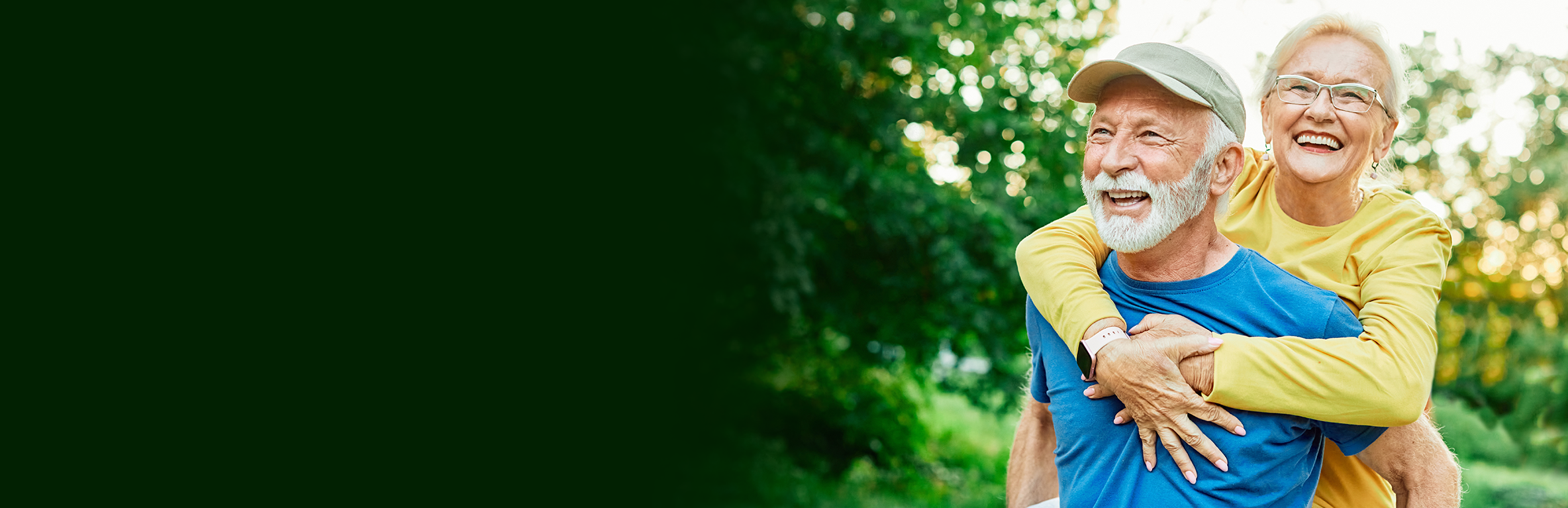 Casal de idoso sorrindo, ele com camisa azul e boné dando carona nas costas para ela que está com blusa amarela e óculos, em ambiente externo com árvores e vegetação ao fundo.