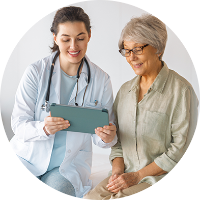 Médica com jaleco branco segurando um tablet e mostrando informações a senhora sentada ao seu lado, em ambiente clínico.