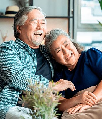 Casal de idosos sentados lado a lado em um sofá, sorrindo. Ao fundo, plantas internas e janelas com vista urbana.