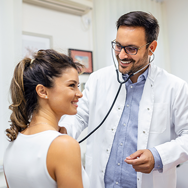 Médico com jaleco branco utilizando estetoscópio para escutar o tórax da paciente sorridente, em ambiente clínico.