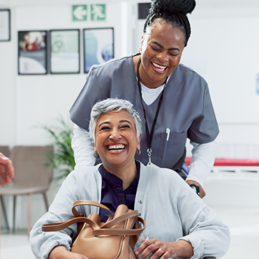 Senhora sentada em cadeira de rodas segurando bolsa marrom e sorrindo, acompanhada por uma enfermeira em pé atrás, também sorridente. Ao fundo, quadros na parede, placa de saída e móveis.