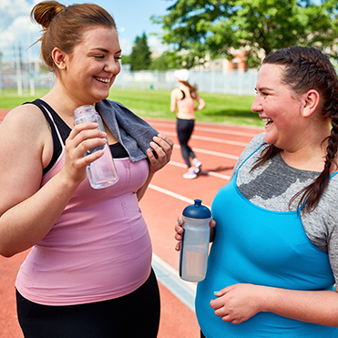Duas mulheres em pé sobre pista de corrida ao ar livre, segurando garrafas de água e conversando. Uma veste regata rosa com toalha sobre o ombro; a outra, regata azul sobre camiseta cinza. Ao fundo, pessoa correndo e área com árvores, em dia ensolarado.