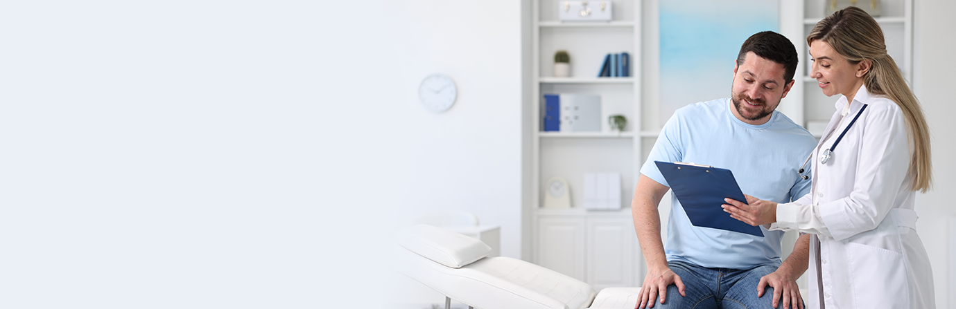 Homem sentado em uma maca de exame, vestindo camisa azul clara e jeans, conversa com a médica, vestida com jaleco branco que segura uma prancheta. Ao fundo, estante com livros, plantas e objetos decorativos, além de um relógio na parede.