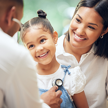 Menina sorridente sendo examinada por um médico com estetoscópio, acompanhada por uma mulher em ambiente médico acolhedor. Imagem representa cuidado pediátrico, confiança no atendimento e saúde infantil.