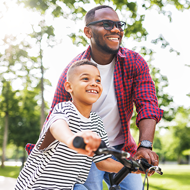 Um homem adulto e um menino sorrindo e andando de bicicleta em um parque arborizado durante dia ensolarado. Companheirismo, lazer ao ar livre e estilo de vida saudável em ambiente natural.