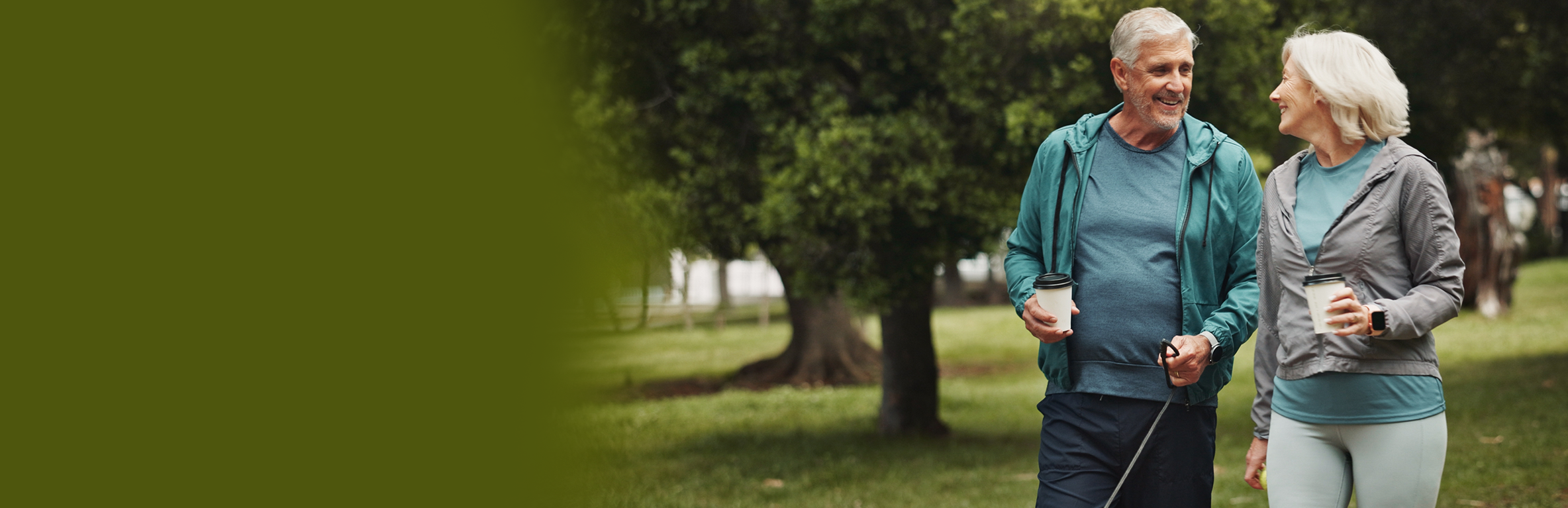 Casal caminhando em parque arborizado, vestindo roupas esportivas casuais e segurando copos de café, em ambiente com grama e árvores verdes.