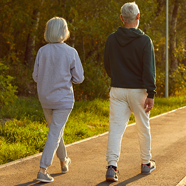 Casal caminhando em trilha pavimentada cercada por vegetação, vestindo roupas confortáveis para atividade física, com iluminação suave indicando manhã ou fim de tarde.
